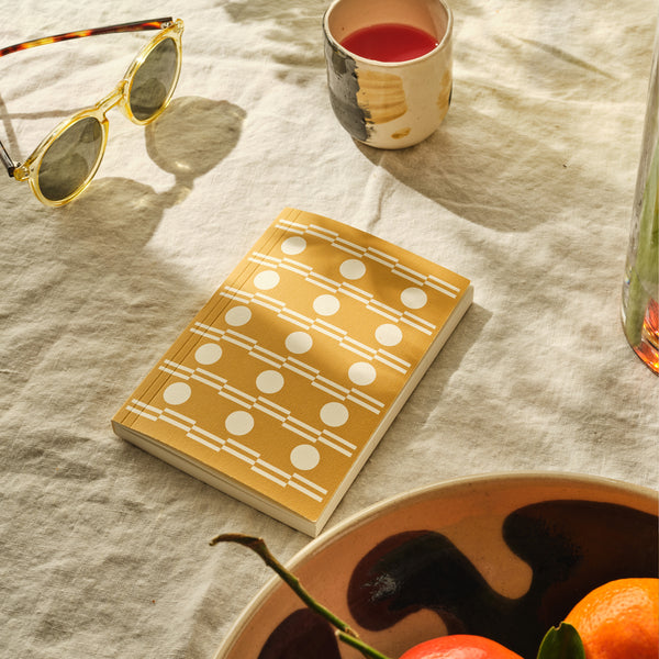 Notebook with geometric pattern on a tablecloth with a bowl of fruit and sunglasses.